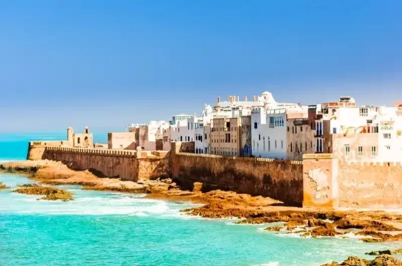 Historic coastal city with white buildings against a vibrant blue sky. Waves crash against the rocky shore beside ancient stone walls, evoking tranquility.