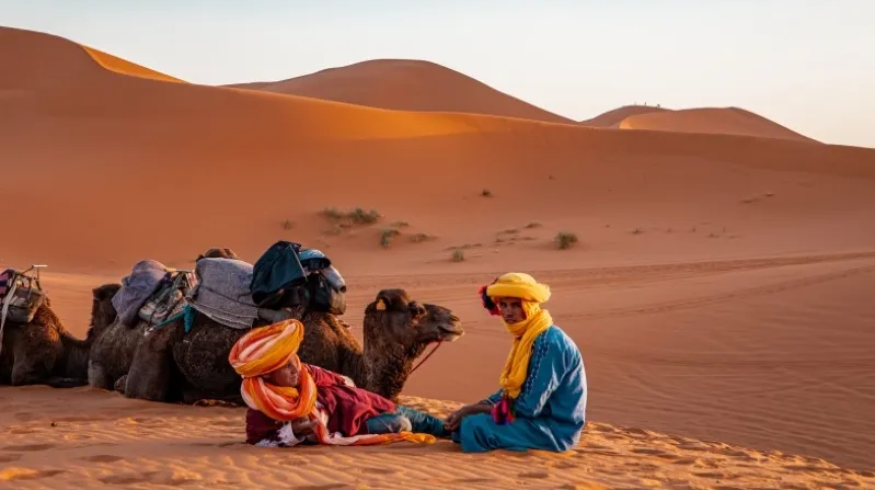 Two people in vibrant clothing sit on the sand beside camels, showcasing a colorful scene in Morocco.