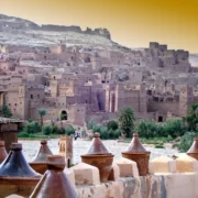 Ancient desert fortress with earthen buildings against a golden sky, surrounded by palm trees. Foreground features clay pots on a stone wall.