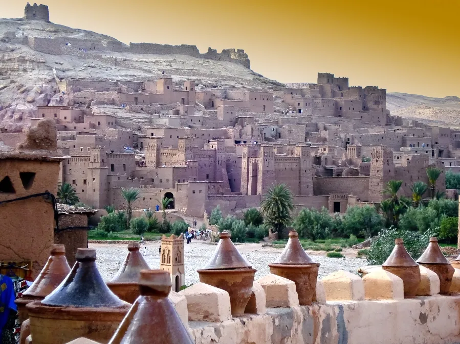 Ancient desert fortress with earthen buildings against a golden sky, surrounded by palm trees. Foreground features clay pots on a stone wall.