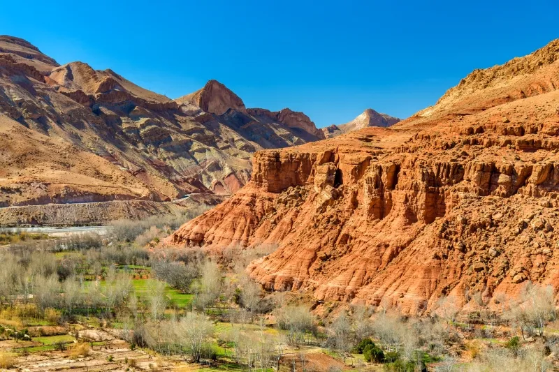 Red cliffs of the Karakoram Mountains rise dramatically in a valley, showcasing the rugged beauty of the Moroccan Atlas Mountains.