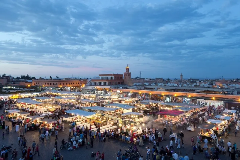 Bustling evening market in Marrakech with glowing stalls and a lively crowd. The sky is dusk blue, adding to the vibrant, energetic atmosphere.Between Marrakech Things to See.