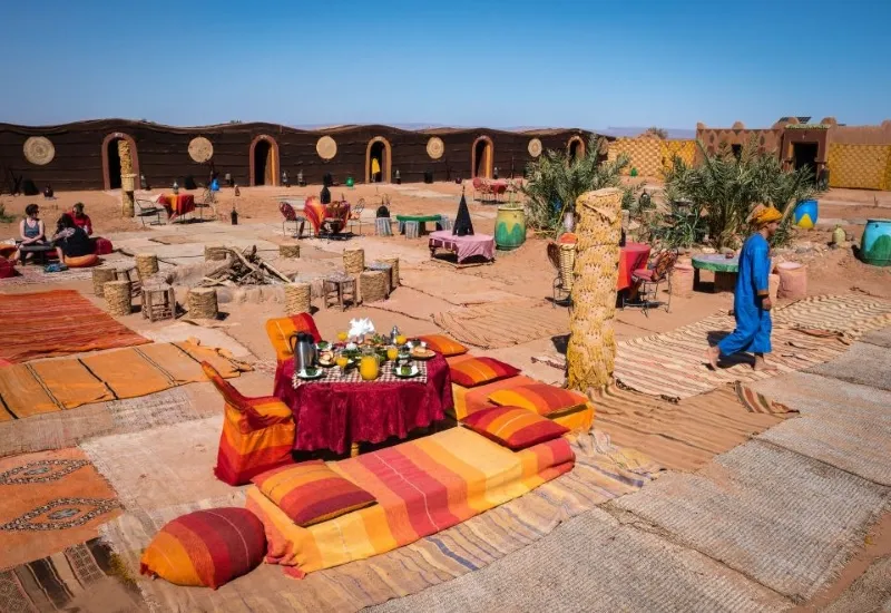Marrakech Desert Tours Desert camp scene with vibrant textiles covering the ground. A table set with food and drinks is in the foreground. People are seated, and a person in blue walks by.