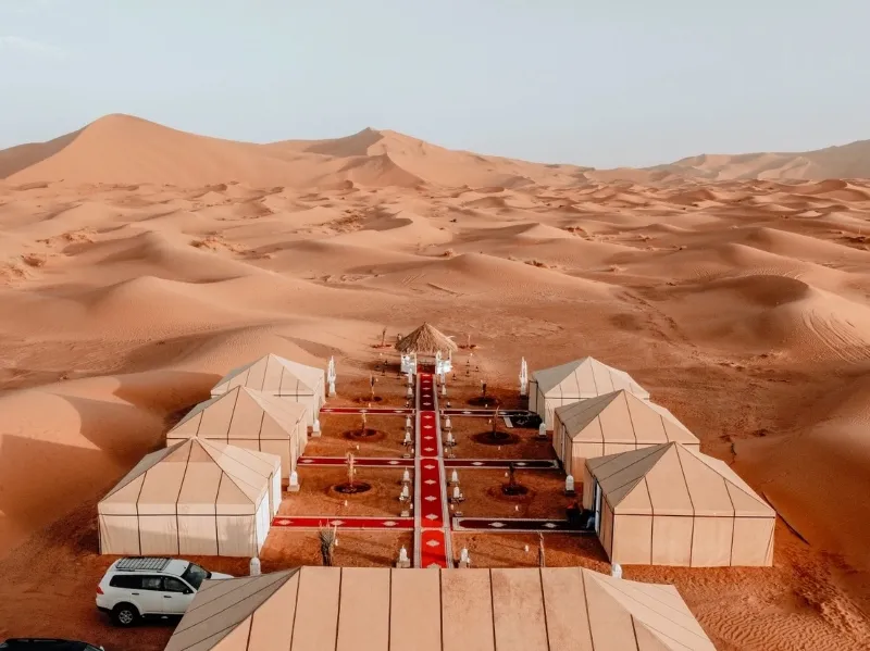 Marrakech Desert Tours (3) Tents with a red carpet path in a desert camp, surrounded by vast, serene sand dunes under a clear sky, creating a peaceful and exotic atmosphere.