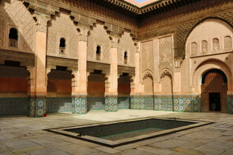 Ornate courtyard of a historic building, featuring intricate tilework, carved archways, and a central rectangular fountain pool under a vibrant blue sky.