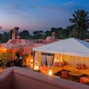 Rooftop patio with a white canopy, warm lighting, orange cushions, and lanterns. Palm trees and sunset sky create a tranquil, exotic ambiance.