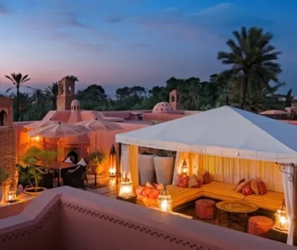 Rooftop patio with a white canopy, warm lighting, orange cushions, and lanterns. Palm trees and sunset sky create a tranquil, exotic ambiance.