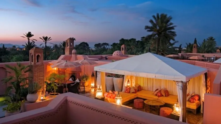 Rooftop patio with a white canopy, warm lighting, orange cushions, and lanterns. Palm trees and sunset sky create a tranquil, exotic ambiance.