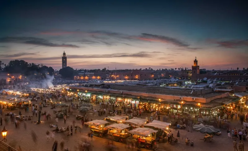 the Magic of Jemaa el Fnaa Bustling evening market in Marrakech with vibrant stalls and blurred crowds under a dusky sky. The skyline features silhouetted minarets.Between best things to do in Marrakech Morocco.